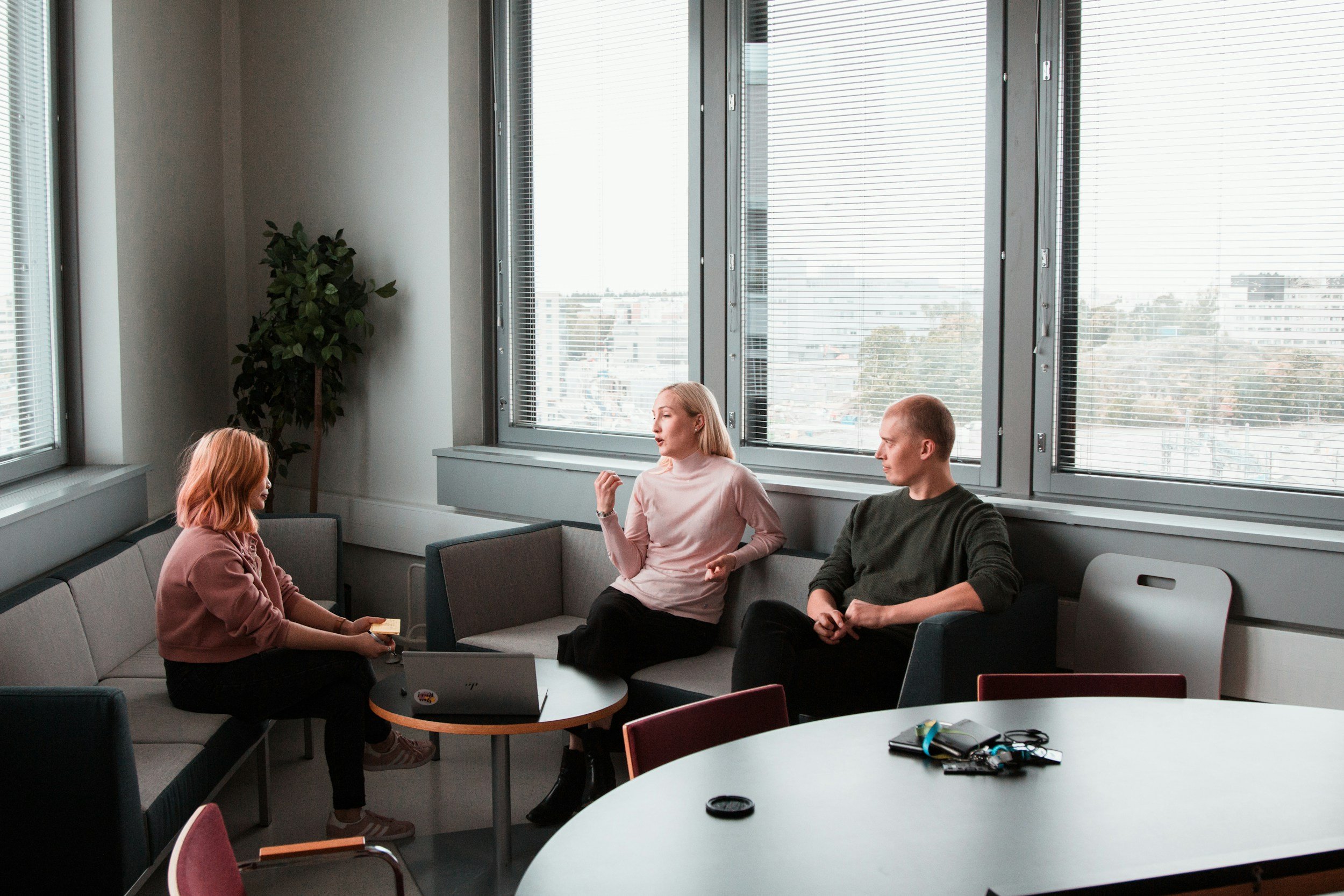Three people having a discussion in a modern office with large windows, a round table, and a laptop.