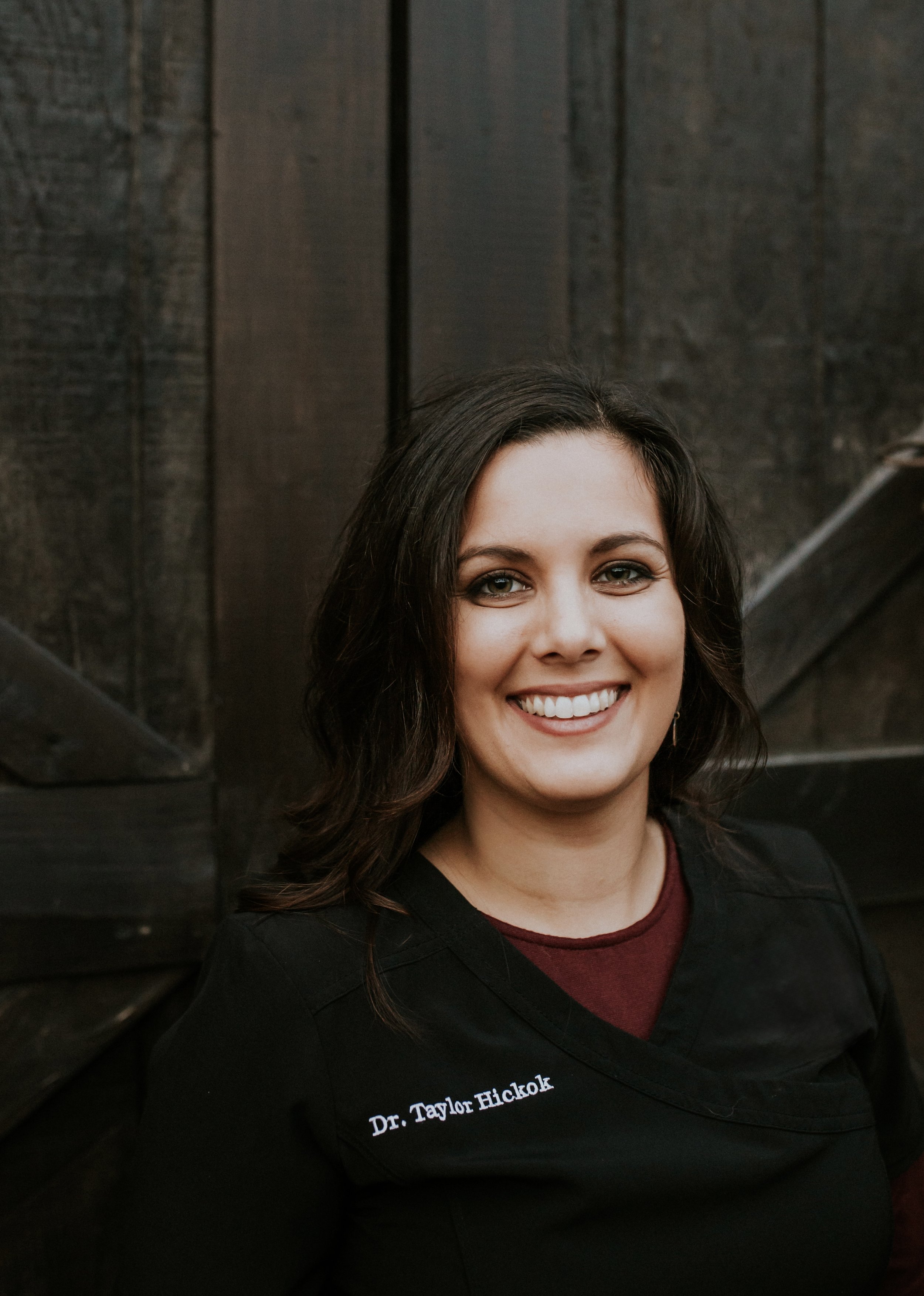 Portrait of Dr. Taylor Hickok, smiling, with dark wavy hair and black scrubs embroidered with her name.