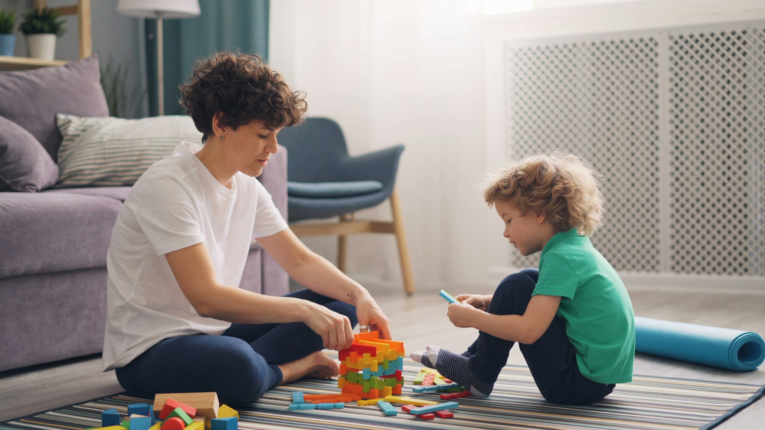 A woman and a young boy playing with colorful building blocks and wooden toys on a striped rug in a living room.