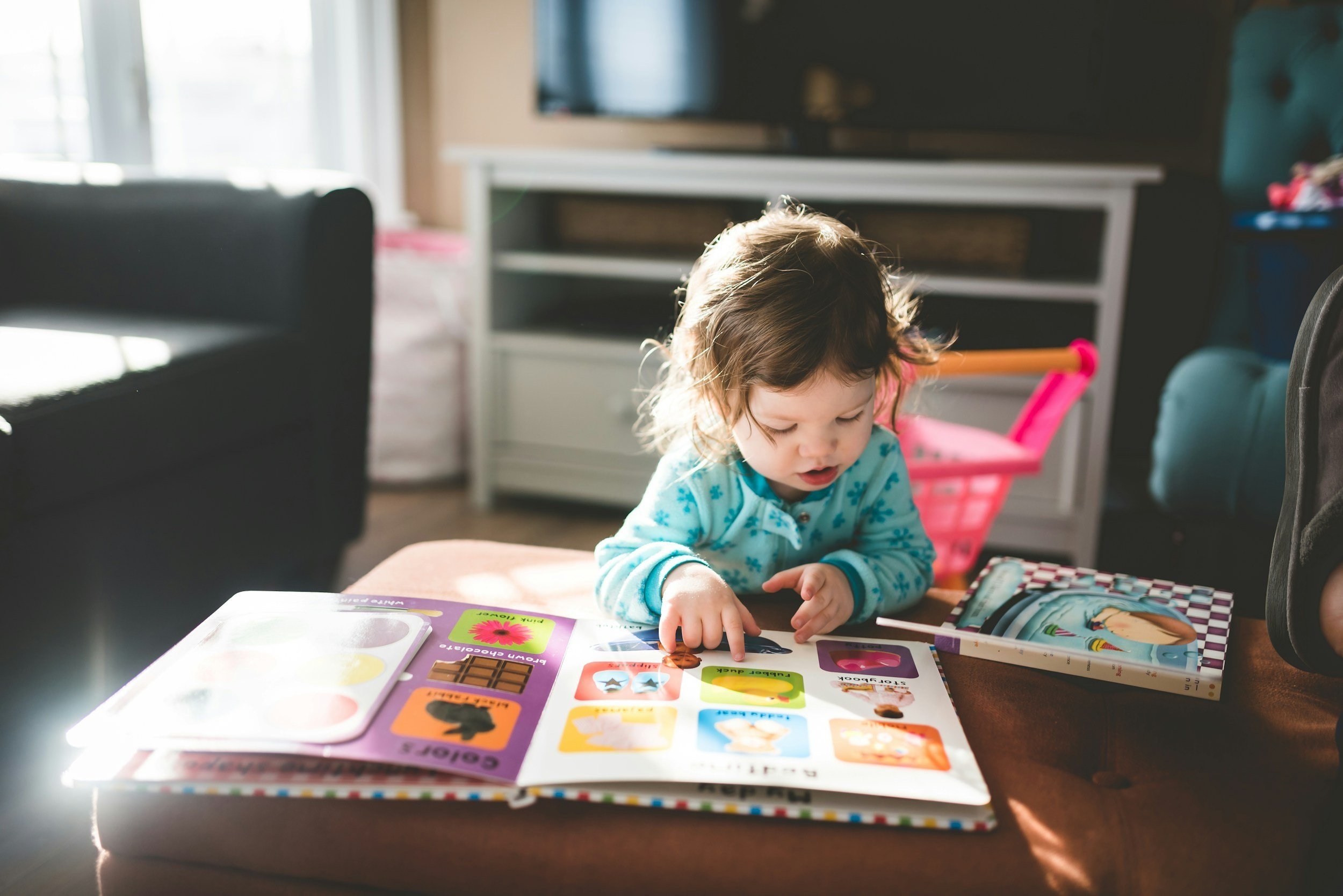 A young child wearing a blue patterned top sits at a table looking at a colorful picture book.